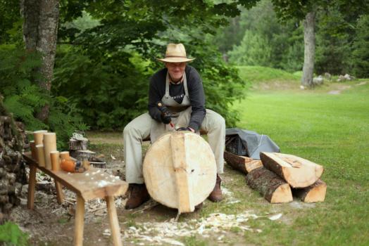 Puutöömeister Meelis Kihulane istub puupakul ja teeb puutööd kaabu peas